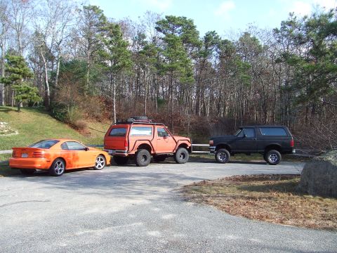 1995 Ford Mustang - An orange 1995 Ford Mustang is parked alongside two Ford Broncos in a wooded area.
