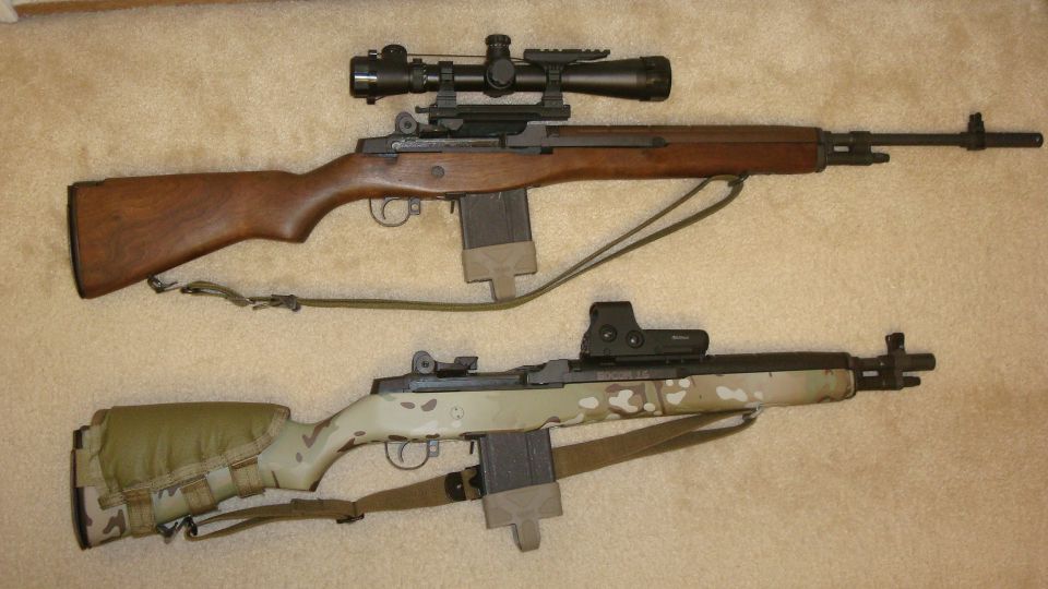 Two rifles on a carpeted surface. One is wood-stocked with a scope. The other has a camouflage finish and a red dot sight
