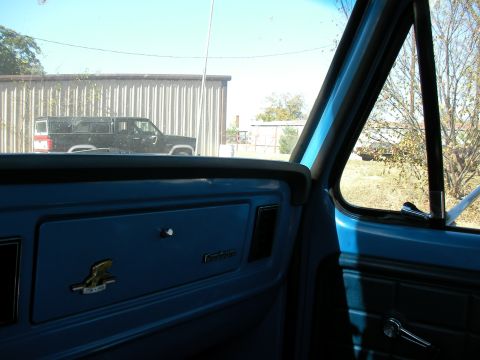 1976 Ford F-150 - Interior view of a blue dashboard with a gold emblem, looking out at a black truck parked outside.