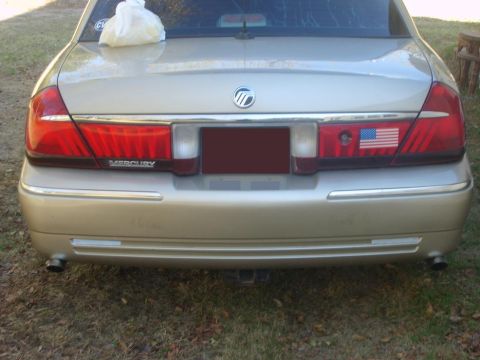 2000 Mercury Grand Marquis - Rear view of a gold 2000 Mercury Grand Marquis, a prominent Mercury emblem and an American flag sticker on the right taillight.