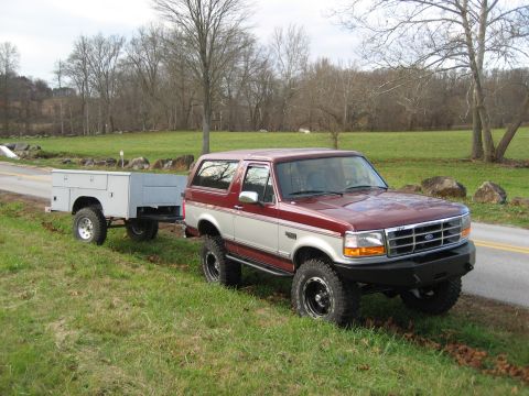 1996 Ford Bronco - A maroon and silver 1996 Ford Bronco is parked on the side of a rural road, attached to a utility trailer.