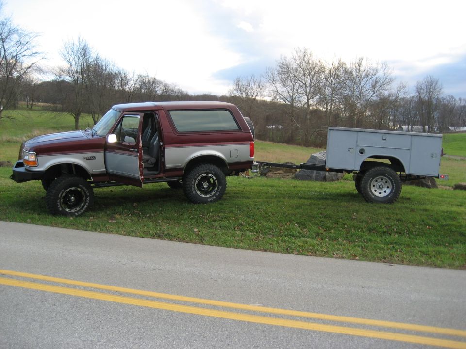 Maroon and silver 1996 Ford Bronco with driver's door open, towing a utility trailer