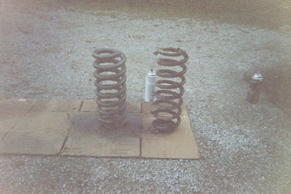 Two coil springs on cardboard over gravel with spray paint cans nearby