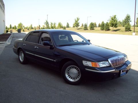 1999 Mercury Grand Marquis - A dark-colored sedan is parked on an asphalt surface with a building and trees in the background.