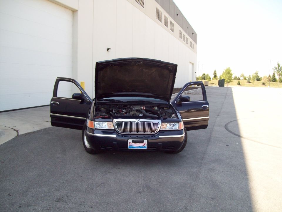 Black sedan with hood and front doors open, parked on asphalt in front of a large building