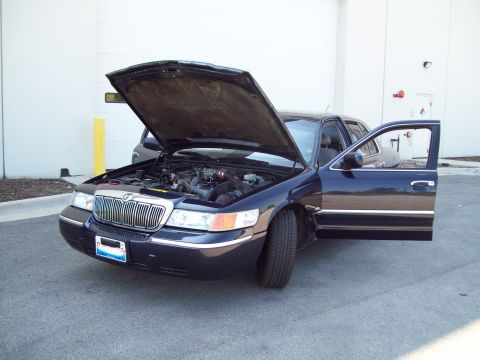 1999 Mercury Grand Marquis - A dark blue 1999 Mercury Grand Marquis is shown with its hood and driver's door open, revealing the engine compartment.