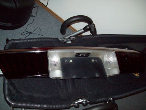 1999 Mercury Grand Marquis - A dark rear license-plate/backup-lamp filler panel from a sedan resting on a black folding chair indoors on gray carpet. The chrome plate s…