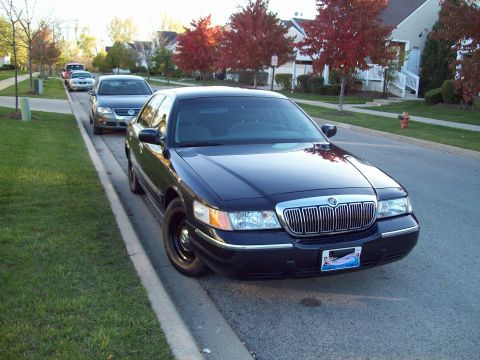 1999 Mercury Grand Marquis - A dark sedan with a chrome grille is parked curbside on a residential street.