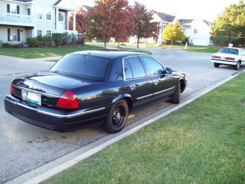 1999 Mercury Grand Marquis - Rear three-quarter view of a black full-size Mercury sedan with steel wheels parked at a residential curb, houses and autumn trees behind.
