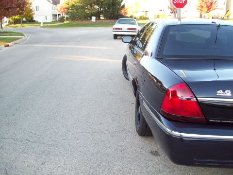 1999 Mercury Grand Marquis - A dark sedan is parked on a residential street, with another white car visible in the background.