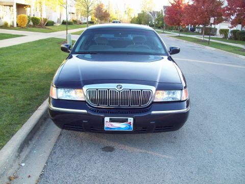 1999 Mercury Grand Marquis - A dark blue 1999 Mercury Grand Marquis is parked curbside on a street with houses and trees in the background.