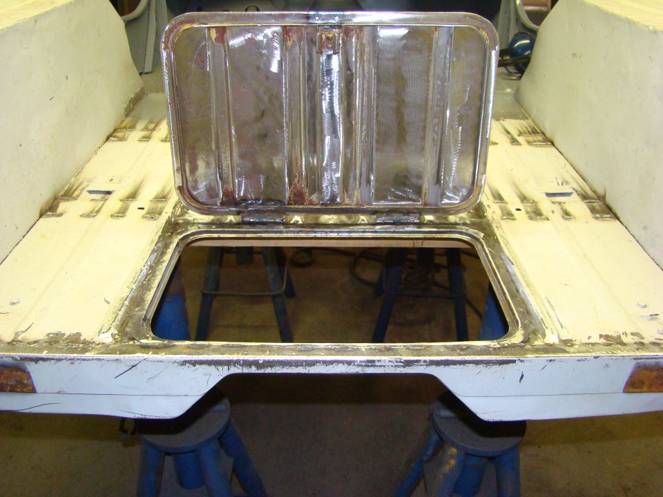 White vehicle body with an open metal access panel revealing a dark interior. The panel is hinged and shows signs of rust