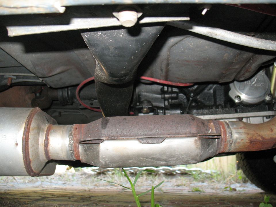 Underside view of a vehicle's exhaust pipe and catalytic converter, showing rust and some wiring