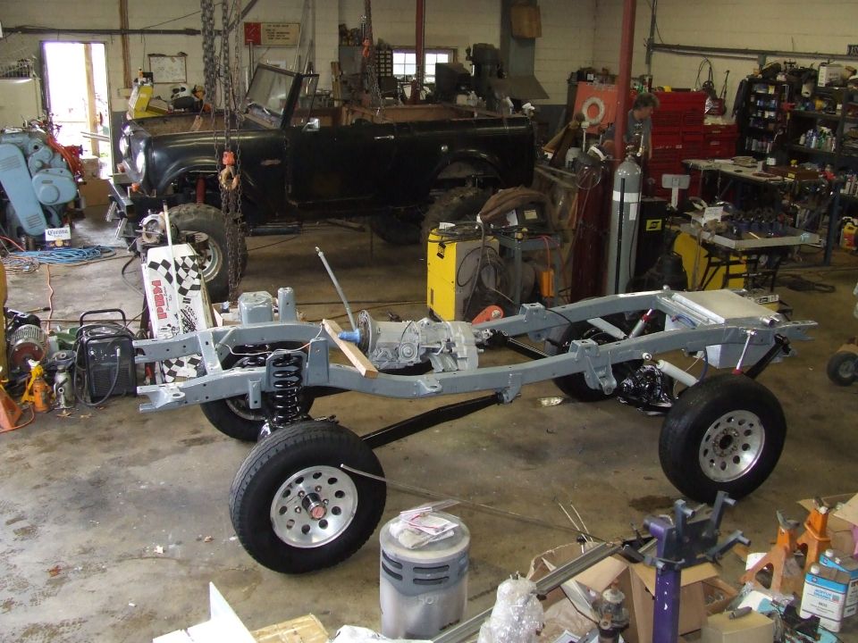 Vehicle frame and drivetrain assembly in a workshop, with a black SUV body in the background