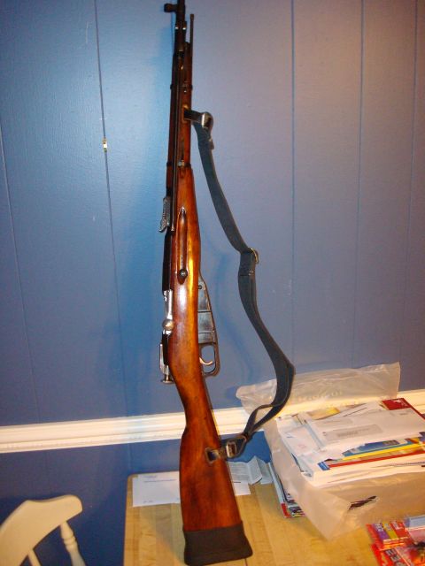1977 Ford F-100 - A rifle with a wooden stock and a black sling is shown standing upright against a blue wall, with a stack of mail in the background.