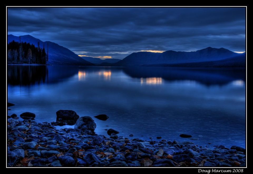 Rocky shore of a calm lake with mountains in the background under a cloudy, dark blue sky. Reflections of the sky and distant lights are visible on the water