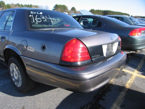 2001 Ford Crown Victoria - A dark gray 2001 Ford Crown Victoria is parked in a lot with other vehicles, showing its rear quarter panel and a dented bumper.