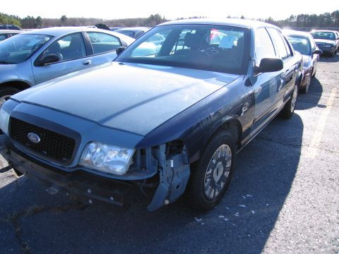 2001 Ford Crown Victoria - A blue Ford Crown Victoria with missing front bumper and headlight sits in a car lot.