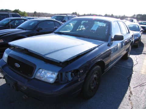 2001 Ford Crown Victoria - A blue 2001 Ford Crown Victoria sits in a junkyard with its hood ajar and one headlight missing.