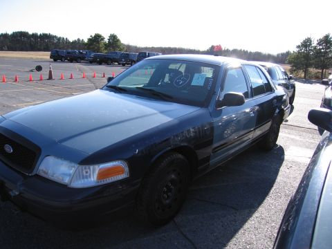 2001 Ford Crown Victoria - A dark blue 2001 Ford Crown Victoria sedan is parked outdoors with other vehicles. Paper tags are visible on the windshield.
