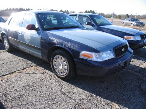 2001 Ford Crown Victoria - Two police cruisers are parked on cracked asphalt under a bright sky. The front vehicle has 'STATE POLICE' written on the hood.