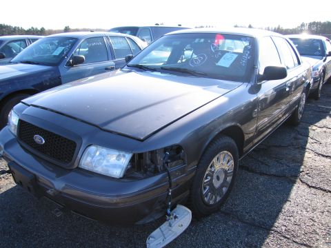 2001 Ford Crown Victoria - A dark gray 2001 Ford Crown Victoria is parked in a lot with other vehicles, showing damage to the front passenger side headlight.