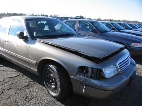 2001 Ford Crown Victoria - A row of gray sedans, including a 2001 Ford Crown Victoria with its hood ajar and a missing headlight, are parked outdoors.