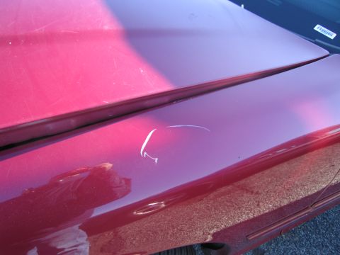 2001 Ford Crown Victoria - A close-up shot of a red car's fender reveals a prominent white scratch and other minor imperfections on the paintwork.