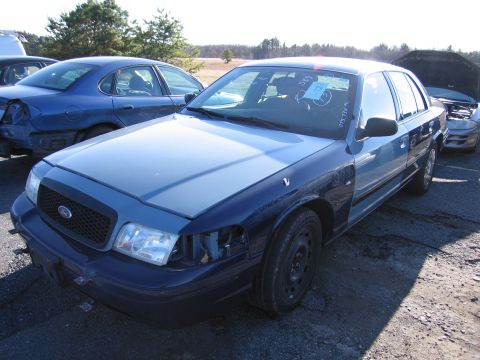 2001 Ford Crown Victoria - A blue Ford Crown Victoria sits in a junkyard, showing signs of damage and repair.