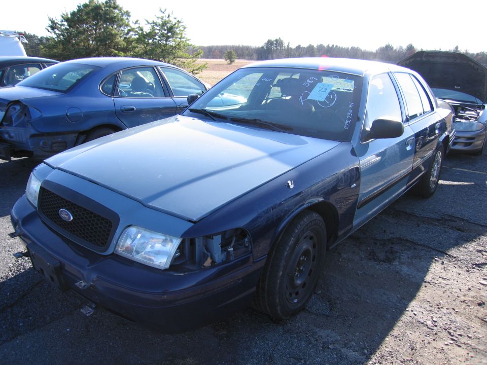 Blue Ford Crown Victoria with a damaged front end and mismatched fenders, parked in a lot with other vehicles