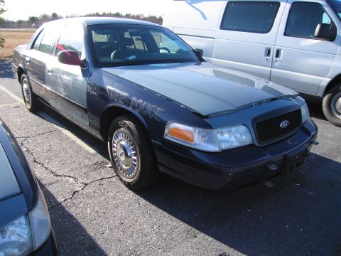 2001 Ford Crown Victoria - A dark blue and silver police car, identified as a 2001 Ford Crown Victoria, is parked next to a white van.