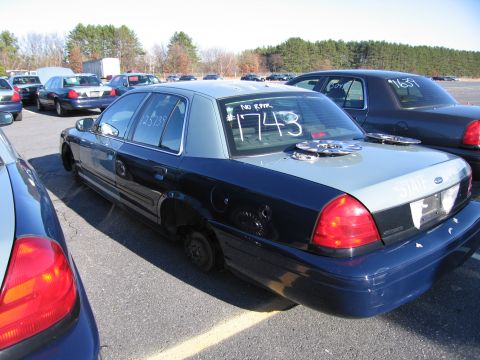 2001 Ford Crown Victoria - A dark blue and grey 2001 Ford Crown Victoria sits in a lot with other vehicles, missing its wheels and with hubcaps on the trunk.