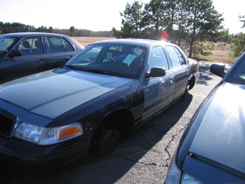 2001 Ford Crown Victoria - A blue Ford Crown Victoria, missing its front wheels, sits in a lot next to other vehicles.