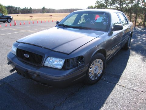 2001 Ford Crown Victoria - A dark gray 2001 Ford Crown Victoria is parked on a cracked asphalt surface with its hood and one headlight missing.