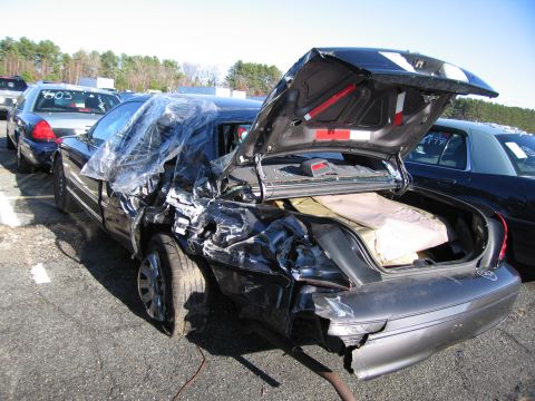 2001 Ford Crown Victoria - A severely damaged sedan sits in a lot with its trunk open, covered in plastic sheeting.