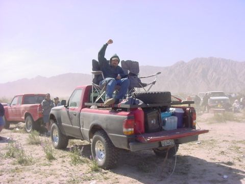 1984 Ford Bronco - A man celebrates from atop a loaded pickup truck in a dusty, outdoor gathering with other vehicles.