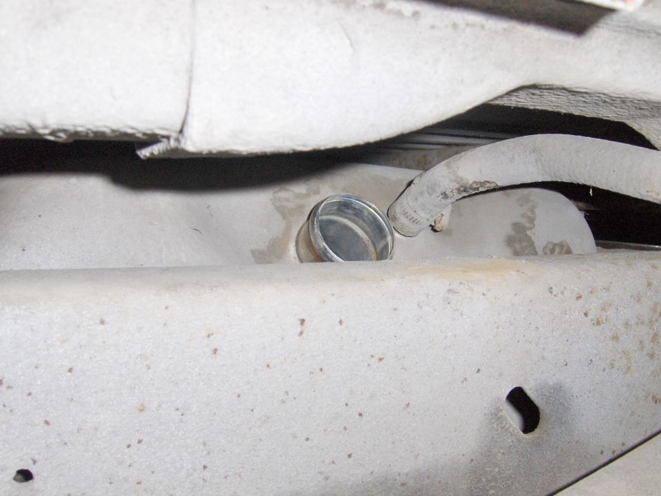 Underside vehicle component with metal filler cap and rubber hose, coated in gray dust.