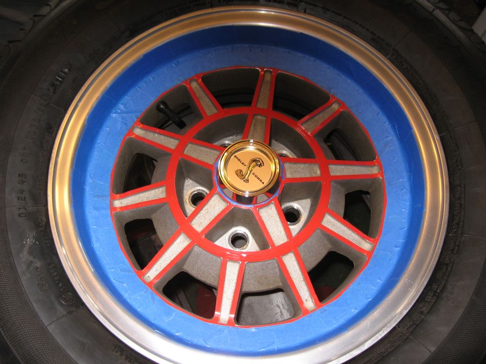Close-up of a car wheel with blue painter's tape, red and silver spokes, and a gold Shelby Cobra center cap