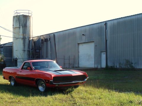 1970 Ford Ranchero - A red 1970 Ford Ranchero is parked on grass in front of an industrial building.