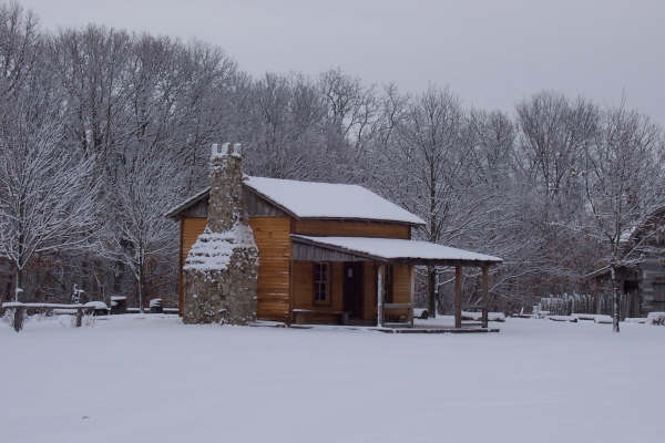 Log cabin with stone chimney and covered porch in deep snow among bare trees