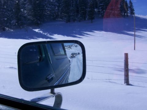 1979 Ford Bronco - A convoy of vehicles travels down a snow-covered road, seen through the side mirror of a green vehicle.
