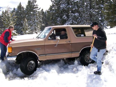 1979 Ford Bronco - A person pushes the front of a stuck Ford Bronco while another digs snow away from the tires.