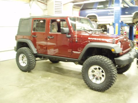 1978 Ford Bronco - Red Jeep Wrangler Rubicon with large tires and custom wheels is parked inside a garage.
