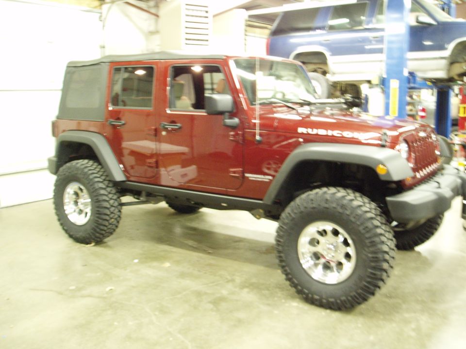 Red Jeep Wrangler Rubicon with aftermarket wheels and tires in a garage. Another vehicle is visible on a lift in the background