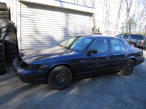 1991 Ford LTD Crown Victoria - A dark blue sedan is parked outside a garage with a stack of tires nearby.