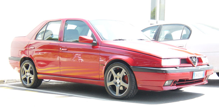 Red Alfa Romeo 155 sedan with dark alloy wheels, front three-quarter view in a parking lot