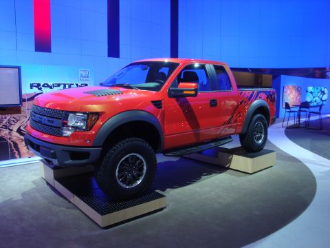 1993 Ford Taurus - A red Ford F-150 Raptor pickup truck is displayed at an auto show, with "RAPTOR" text visible on a backdrop.