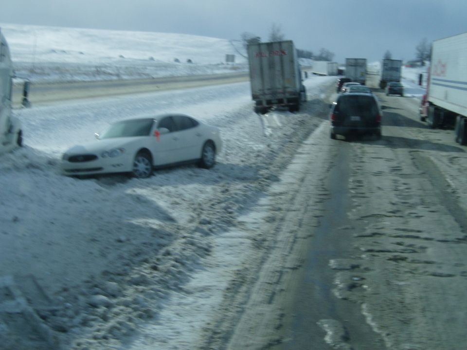 White sedan partially off the road in snow, with trucks and other vehicles on a slushy highway