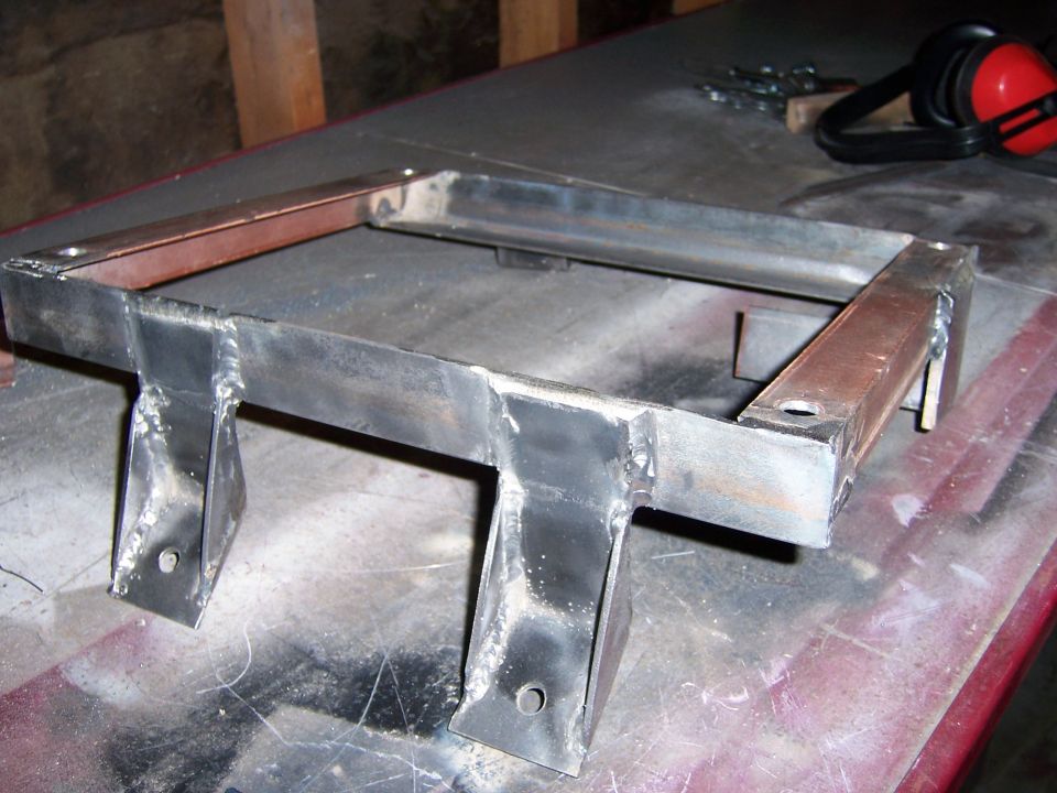 Welded metal bracket with four mounting tabs, resting on a workbench