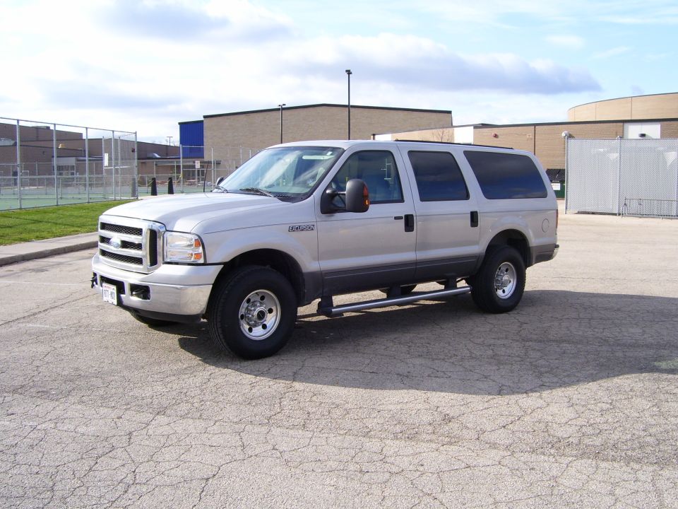 Silver Ford Excursion parked on cracked asphalt, with a chain-link fence and buildings in the background